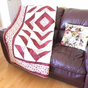 Può includere: Un trapunta rosso e bianco con un motivo geometrico è drappeggiato su un divano in pelle bordeaux. La trapunta ha uno sfondo bianco con forme geometriche rosse e un bordo rosso con motivi di foglie d'acero bianche.