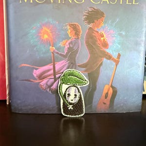 May include: An embroidered patch featuring a black, ghost-like figure with a white mask and purple facial markings, holding a green leaf over its head. The patch has a white border and is positioned in front of a book cover titled Howl's Moving Castle.