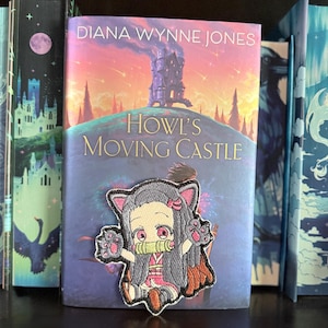 May include: An embroidered patch featuring a chibi-style character with long dark hair, pink eyes, and cat ears, wearing a pink and white outfit with a green bamboo-style muzzle, positioned on the cover of the book Howl's Moving Castle.