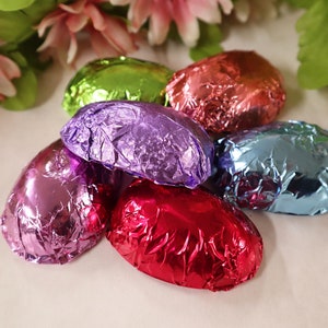 May include: Six foil-wrapped chocolate eggs in various colors, including purple, green, red, pink, and blue. The eggs are arranged in a cluster on a white surface.