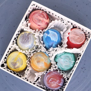 May include: A white box filled with nine assorted chocolates. The chocolates are all different colors and have a swirl pattern. The chocolates are arranged in a circle.