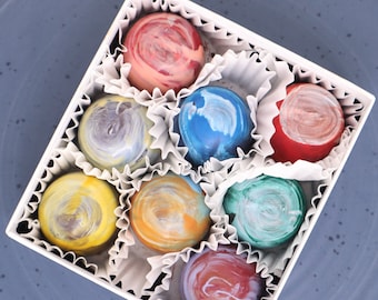 Puede incluir: Una caja blanca llena de nueve chocolates surtidos. Los chocolates son de diferentes colores y tienen un patrón de espiral. Los chocolates están dispuestos en círculo.