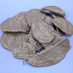 May include: A pile of chocolate-covered potato chips on a light purple surface. The chips are a light brown color and have a wavy texture. The image is well-lit and the chips are in focus.