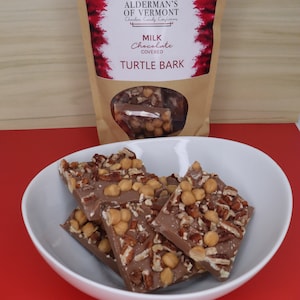 May include: A white bowl filled with milk chocolate turtle bark. The bark is covered in pecans and caramel pieces. The bowl is sitting on a red surface. The bag of bark is in the background with the brand name "Alderman's of Vermont" and the text "Milk Chocolate Covered Turtle Bark".