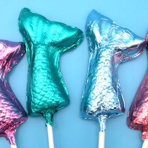 May include: Four mermaid tail-shaped lollipops in shades of pink, teal, and blue, each on a white stick. The lollipops have a metallic, foil-like appearance and are arranged against a light blue background.