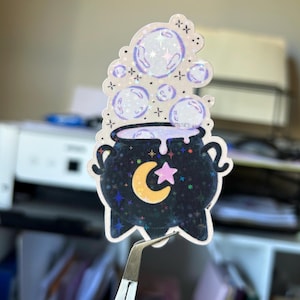 May include: A holographic sticker depicting a black cauldron with a crescent moon and star design. Bubbles with stars and cross accents float above the cauldron. The sticker is held by tweezers.
