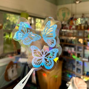 May include: A collection of three butterfly stickers. Each butterfly is light blue with detailed wing designs. The stickers have a holographic, iridescent effect, reflecting various colours. The stickers are held by silver tweezers.