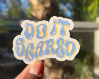 Op de afbeelding: Een blauwe en gele sticker met de tekst "Do It Scared" in een bubbelvormig lettertype.