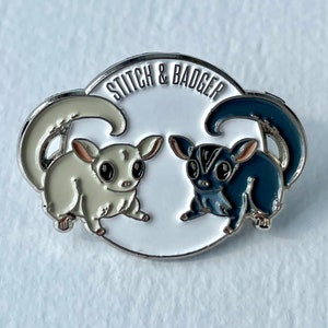 May include: A white enamel pin with a silver border featuring two sugar gliders facing each other. The sugar glider on the left is white and the sugar glider on the right is gray. The text "Stitch & Badger" is written in a silver font around the top of the pin.
