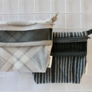 Pode incluir: Duas bolsas penduradas com janelas de malha e fechos de correr. A bolsa à esquerda é branca com um padrão xadrez cinzento e a bolsa à direita é cinzenta com riscas pretas. Ambas as bolsas têm correias pretas.