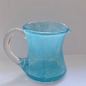 Puede incluir: Una jarra de vidrio azul claro con un asa curva y un pico vertedor. La jarra tiene una superficie ligeramente texturizada y una base redondeada. El asa es de vidrio transparente y la jarra está sobre un fondo blanco.