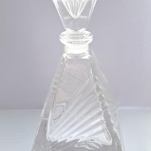 May include: Clear glass decanter with a geometric design. The decanter has a triangular shape with a square stopper. The stopper has a diamond-shaped top. The glass has a textured, fan-like pattern.