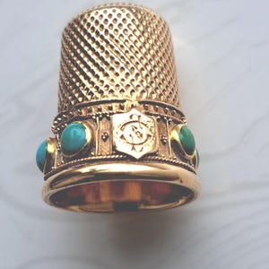 Puede incluir: Un dedal dorado con superficie texturizada y banda decorativa. La banda presenta piedras de color turquesa y un emblema central. El dedal está diseñado para coser y manualidades.