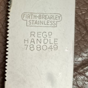 May include: Close-up of a silver knife blade with a serrated edge. The blade is stamped with the text "Firth-Brearley Stainless" and "Reg'd Handle 788049".