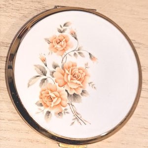 May include: A round, vintage compact mirror with a gold-tone metal frame. The white surface features a floral design of peach-colored roses and gray leaves. The compact is open, revealing the mirror inside.