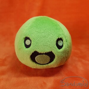 Può includere: Un peluche verde lime a forma di creatura rotonda con occhi neri ricamati e una bocca sorridente. Il giocattolo ha una consistenza morbida e soffice ed è posto su uno sfondo arancione. Un design carino e stravagante.