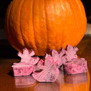 Puede incluir: Una gran calabaza de color naranja brillante con un tallo azul oscuro se encuentra detrás de varios recortes de fantasmas de madera rosa. Los fantasmas son pequeños y tienen un aspecto rústico e inacabado. La escena probablemente esté relacionada con la decoración de Halloween o otoño.