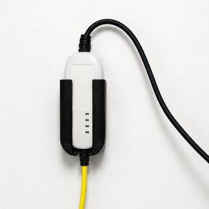 Op de afbeelding: Een wit en zwart elektrisch voertuig oplader met een zwarte kabel en een gele kabel. De oplader heeft een rechthoekige vorm met vier kleine indicatielampjes. De achtergrond is effen wit.