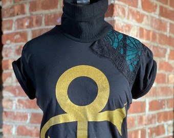 May include: Black short-sleeved shirt with a gold Prince symbol graphic and a teal lace detail on the shoulder.