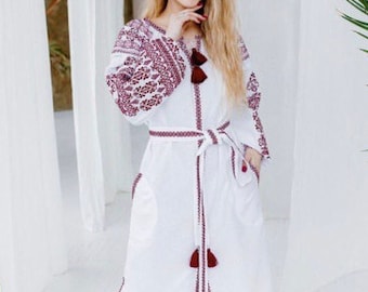 May include: White linen dress with long sleeves and a burgundy embroidered design. The dress has a belt and a front slit.