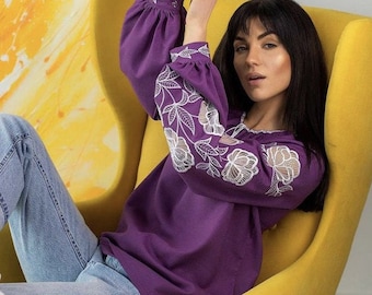 Puede incluir: Una mujer lleva una blusa morada con diseños florales bordados en blanco. Está sentada en un sillón amarillo.