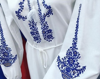 May include: White embroidered dress with blue floral patterns. The dress has long sleeves with a gathered cuff and a white tie at the waist.