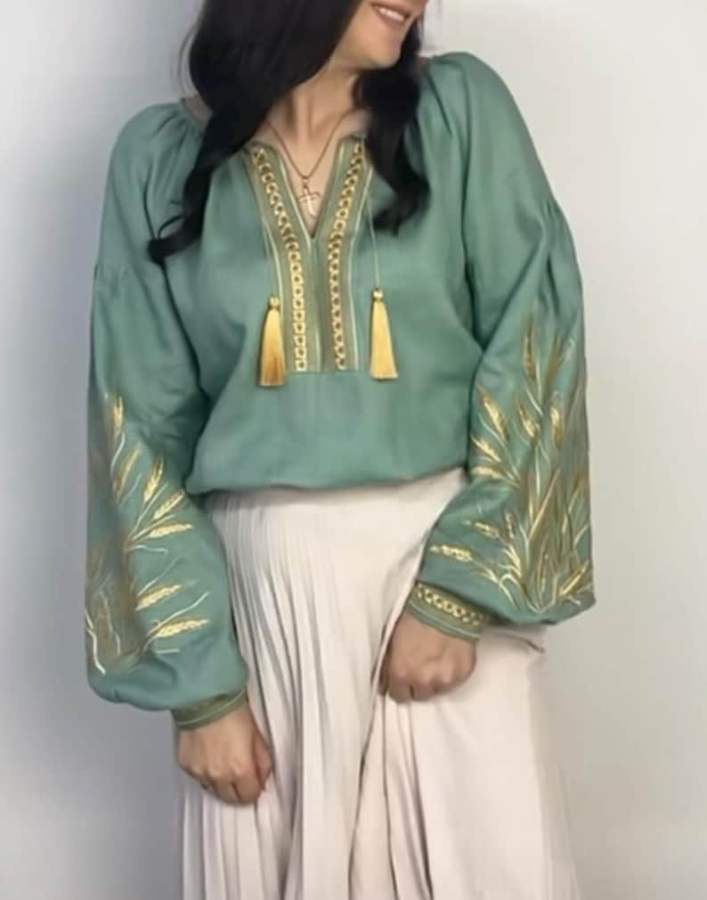 May include: A sage green linen blouse with gold embroidery of wheat stalks on the sleeves and neckline. The blouse has a V-neck and a gathered neckline with tassels.
