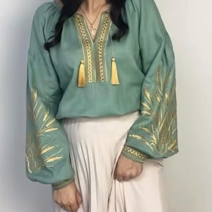 May include: A sage green linen blouse with gold embroidery of wheat stalks on the sleeves and neckline. The blouse has a V-neck and a gathered neckline with tassels.