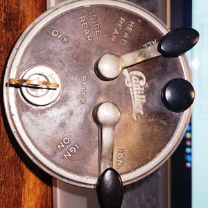 May include: Vintage Cadillac dashboard control panel with multiple levers and a key. The panel is circular, with text indicating light and ignition controls. The levers have black knobs. The panel is made of metal and shows signs of age.