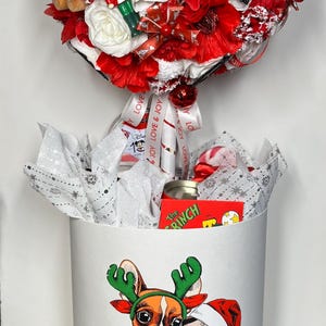 May include: A Christmas-themed gift basket with a decorative topiary of red and white flowers, ornaments, and a nutcracker. The white cylindrical base features illustrations of dogs in Christmas attire, with the text "MERRY CHRISTMAS AND HAPPY NEW YEAR".
