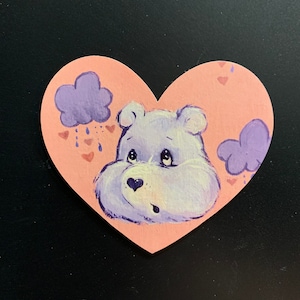 May include: A hand-painted heart-shaped wooden ornament featuring a purple Care Bear with a sad expression. The background is pink with purple clouds and pink hearts.