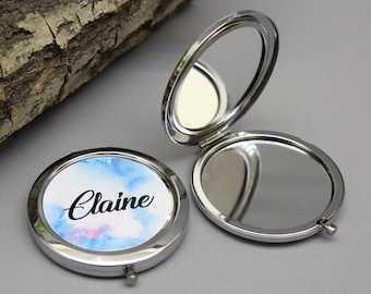 Peut inclure: Deux miroirs compacts argentés ouverts. L'un présente un motif aquarelle aux teintes bleues et roses avec le nom "Claire" en cursive. L'autre miroir est ouvert, reflétant l'environnement. Les miroirs sont sur une surface grise.