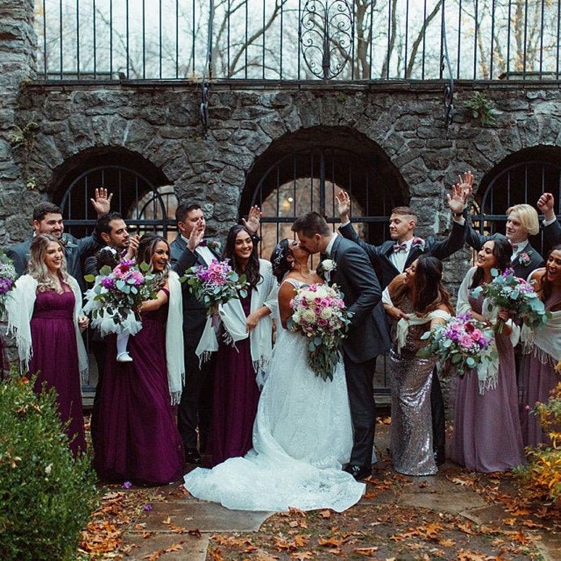 May include: A wedding party poses for a photo in front of a stone archway. The bride and groom are kissing in the center of the group, while the bridesmaids and groomsmen stand around them. The bridesmaids are wearing long, purple dresses, and the groomsmen are wearing black suits. The bride is wearing a white wedding dress with a long train. The wedding party is holding bouquets of flowers.