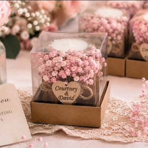 Pu&ograve; includere: Una scatola quadrata in plastica trasparente contenente una candela tea light bianca circondata da fiori di gipsofila rosa. Un'etichetta in legno a forma di cuore &egrave; legata con dello spago, con il testo Courtney & David stampato in nero.