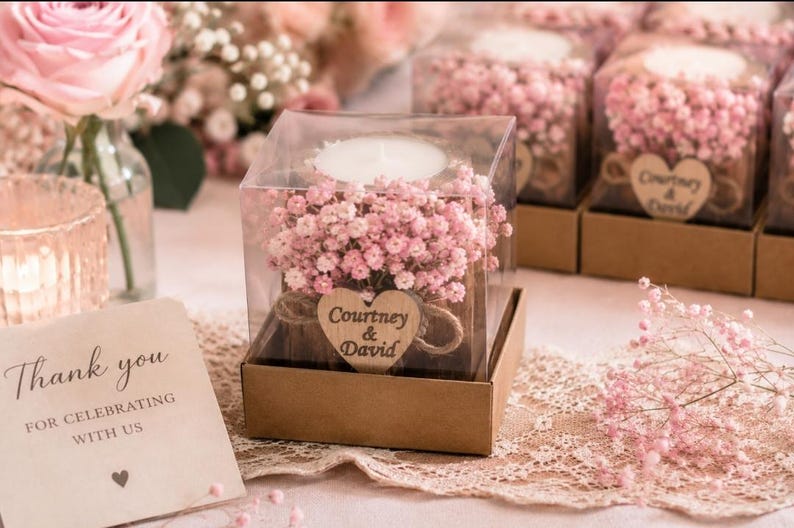 Pu&ograve; includere: Una scatola quadrata in plastica trasparente contenente una candela tea light bianca circondata da fiori di gipsofila rosa. Un'etichetta in legno a forma di cuore &egrave; legata con dello spago, con il testo Courtney & David stampato in nero.