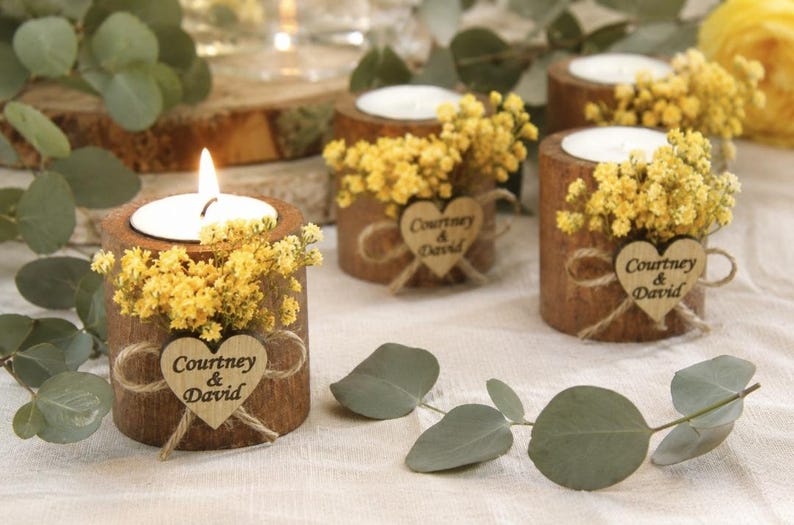 Pu&ograve; includere: Tre portacandele cilindrici in legno con lumini, decorati con fiori gialli e una piccola etichetta in legno a forma di cuore legata con dello spago. Ogni cuore reca l'incisione del testo Courtney & David su un tessuto chiaro.