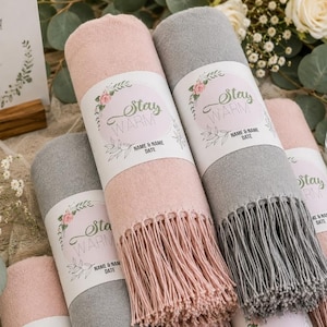 May include: A collection of rolled blankets in soft pink and grey, each featuring a white paper band with the text "Stay WARM" and "NAME & NAME DATE" surrounded by a floral wreath illustration. The blankets have fringed ends and a textured fabric.