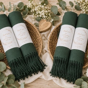 May include: Four dark green rolled scarves with fringed ends, each wrapped in a white paper band featuring the text Thank you for celebrating with us and a floral wreath design. The scarves are arranged in two round woven baskets.