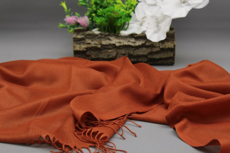 May include: A rust-colored scarf with fringe edges. The scarf is made of a soft, lightweight fabric and is draped over a gray surface.