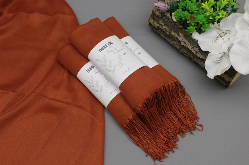May include: Three rust-colored scarves with fringed edges are stacked on a gray surface. Each scarf has a white label attached with the text "Thank You for being my bridesmaid".