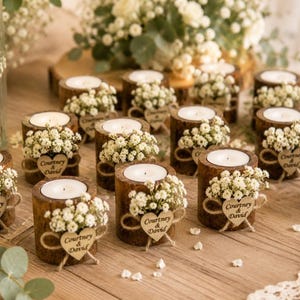 Pu&ograve; includere: Una collezione di portacandele cilindrici in legno, ciascuno dotato di una candelina bianca, un mazzetto di piccoli fiori bianchi e un'etichetta in legno a forma di cuore incisa con il testo Courtney & David, disposti su una superficie in legno.