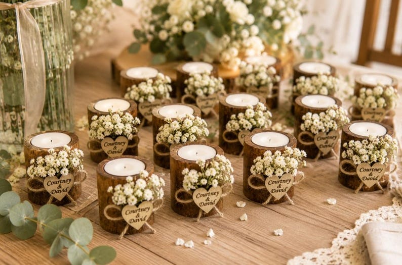 Pu&ograve; includere: Una collezione di portacandele cilindrici in legno, ciascuno dotato di una candelina bianca, un mazzetto di piccoli fiori bianchi e un'etichetta in legno a forma di cuore incisa con il testo Courtney & David, disposti su una superficie in legno.