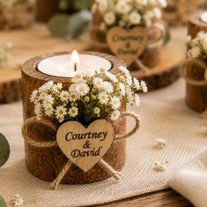 Pu&ograve; includere: Un portacandele cilindrico in legno con un lumino acceso, decorato con fiori bianchi di gipsofila e un'etichetta in legno a forma di cuore legata con dello spago. Il testo Courtney & David &egrave; inciso sull'etichetta in un carattere scuro.