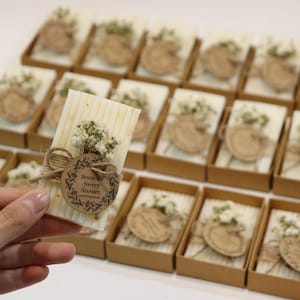May include: A selection of rectangular, off-white favours, each decorated with dried flowers and a wooden tag. The tag reads "Jessica's Sweet Sixteen". The favours are presented in small, brown boxes, ideal for a special occasion.
