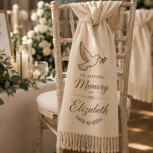 May include: A cream-coloured fabric chair sash with fringe at the bottom, draped over a light wood chair. The sash features a black graphic of a dove holding an olive branch, with the text "IN LOVING Memory OF Elizabeth" and small heart icons.