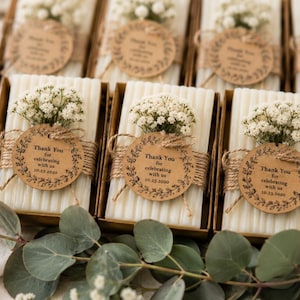 Può includere: Candele rettangolari bianche a coste in scatole marroni individuali, ciascuna decorata con un piccolo bouquet di fiori bianchi di gipsofila e un'etichetta rotonda in carta kraft legata con spago che riporta Thank You for celebrating with us 10.23.2020.