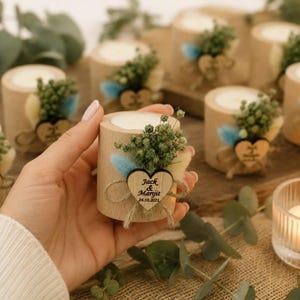 Pu&ograve; includere: Un portacandele cilindrico in legno con una candela in cera bianca, decorato con fogliame verde essiccato, erba secca blu e color crema, e una piccola etichetta in legno a forma di cuore incisa con il testo Jack & Maryit 24.10.2021 legato con spago.