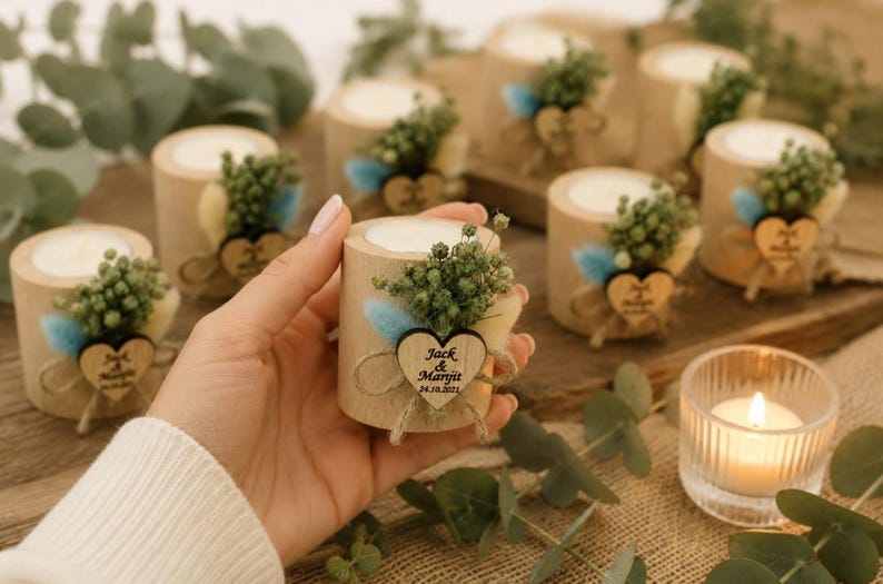 Pu&ograve; includere: Un portacandele cilindrico in legno con una candela in cera bianca, decorato con fogliame verde essiccato, erba secca blu e color crema, e una piccola etichetta in legno a forma di cuore incisa con il testo Jack & Maryit 24.10.2021 legato con spago.