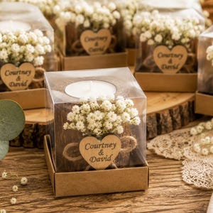 Pu&ograve; includere: Una scatola quadrata in plastica trasparente contenente un portacandele in legno con una candelina bianca. Il supporto &egrave; decorato con fiori bianchi di gipsofila e un'etichetta in legno a forma di cuore con il testo Courtney & David.