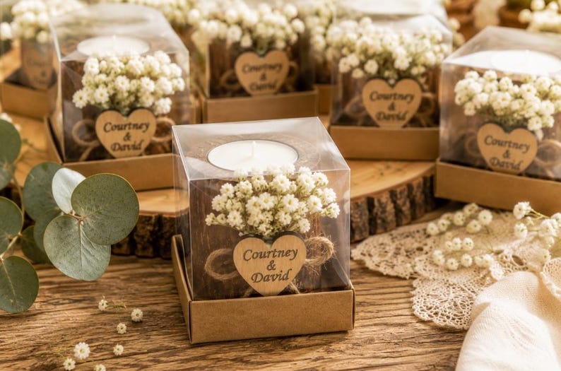 Pu&ograve; includere: Una scatola quadrata in plastica trasparente contenente un portacandele in legno con una candelina bianca. Il supporto &egrave; decorato con fiori bianchi di gipsofila e un'etichetta in legno a forma di cuore con il testo Courtney & David.